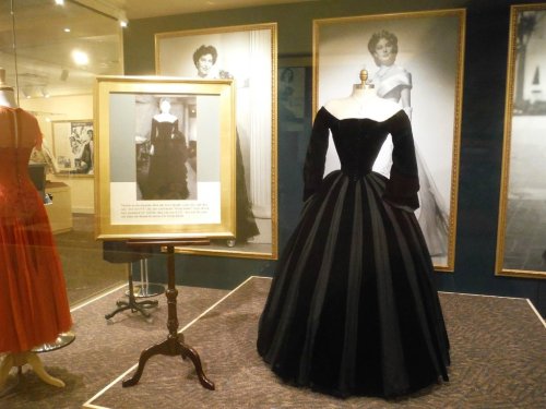 A black evening gown displayed in a museum, surrounded by vintage photographs of elegant dresses.