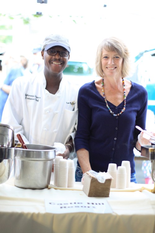 A chef and a woman smile behind a table with cups and food containers at an outdoor event.