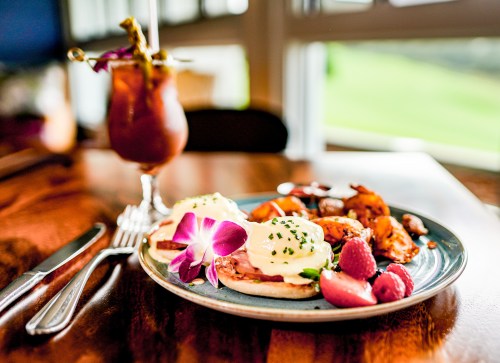 A plate of eggs Benedict with a side of roasted potatoes and a colorful cocktail on a wooden table.