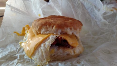 A close-up of a biscuit sandwich with melted cheese, bacon, and a fried egg, wrapped in white paper.