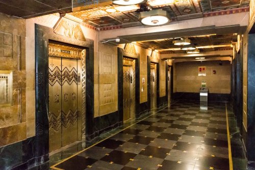 Art Deco hallway with ornate elevator doors, patterned tile floor, and decorative ceiling lights.