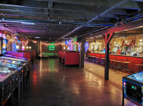 A dimly lit arcade bar with pinball machines, colorful lights, and a long bar in a rustic setting.
