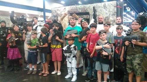 A group of children and adults in costumes, holding toy weapons, posing together at a themed event.