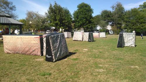A grassy field with several paintball bunkers designed to look like brick walls, surrounded by trees and a clear blue sky.