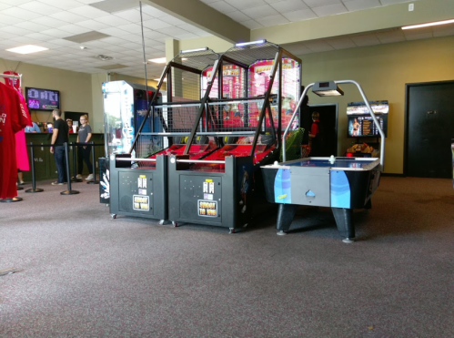 Two arcade games in a game room: a basketball shooting game and an air hockey table, with people in the background.