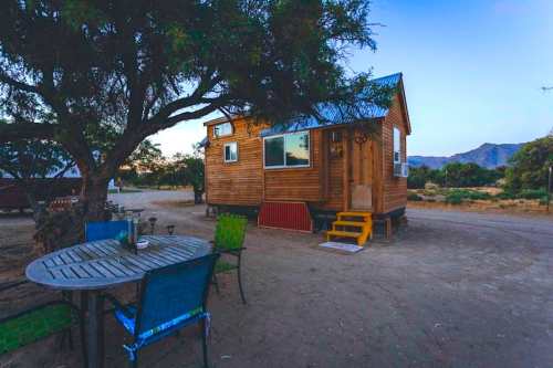 A cozy wooden tiny house beside a table and chairs, set in a serene outdoor landscape with mountains in the background.