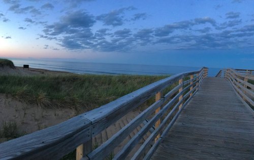 A wooden boardwalk leads to a calm beach at dusk, with gentle waves and a cloudy sky.