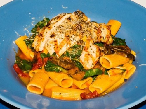 A blue bowl filled with rigatoni pasta, grilled chicken, spinach, and herbs, topped with cheese.