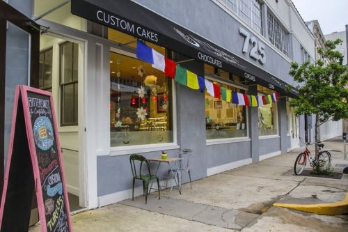 A bakery storefront with colorful flags, outdoor seating, and a chalkboard menu on the sidewalk.
