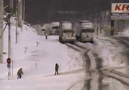 Two trucks on a snowy road with pedestrians walking nearby, surrounded by a winter landscape and a KFC sign in the background.