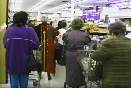 A crowded grocery store aisle with shoppers pushing carts, mostly older women in various outfits.