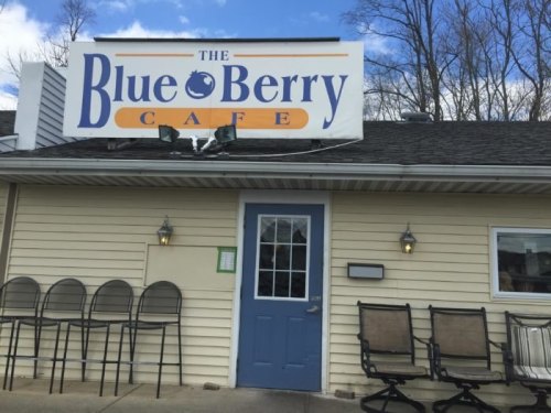 Exterior of The Blue Berry Cafe, featuring a sign, blue door, and stacked chairs outside under a clear sky.