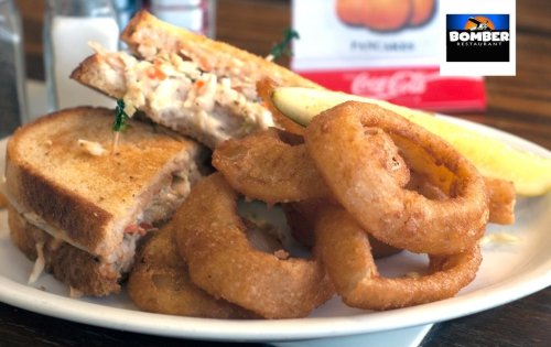 A plate featuring a sandwich with coleslaw and onion rings, accompanied by a lemon wedge. Bomber Restaurant logo in the corner.