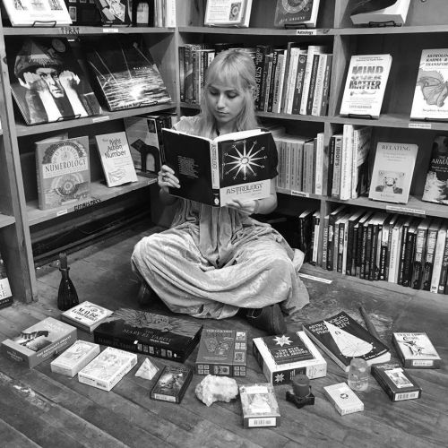 A person sits cross-legged on the floor of a bookstore, reading an astrology book surrounded by tarot cards and crystals.