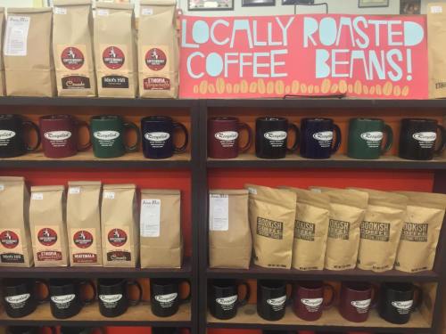 Shelves displaying bags of locally roasted coffee beans and colorful mugs, with a sign highlighting the coffee selection.