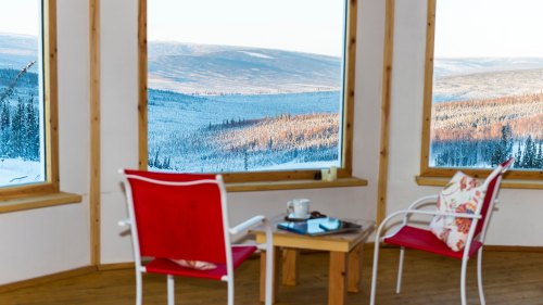 Cozy interior with two red chairs and a table, overlooking a snowy mountain landscape through large windows.