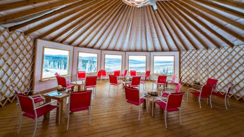 Interior of a yurt with wooden floors, red chairs, and large windows showcasing a snowy landscape outside.