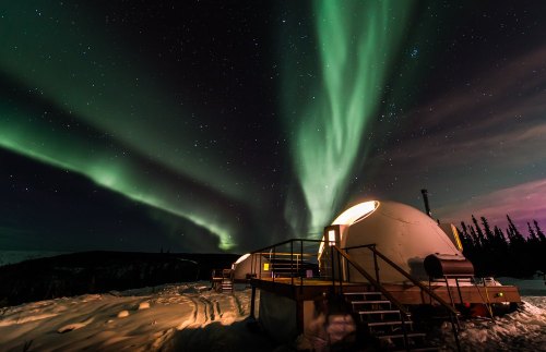 A snowy landscape with futuristic domes under a vibrant display of the Northern Lights in a starry night sky.