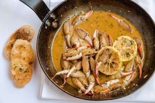 A skillet filled with crab claws in a savory sauce, garnished with lemon slices and served with toasted bread.