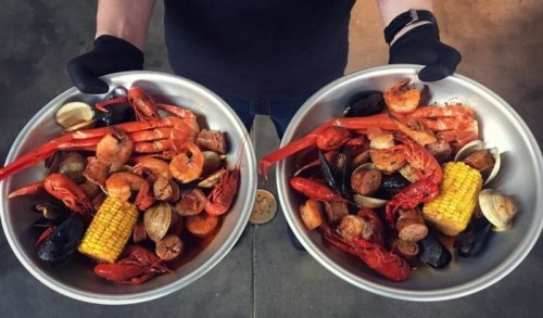 Two large metal bowls filled with a variety of seafood, including shrimp, crawfish, corn, and sausage, held by a person in gloves.