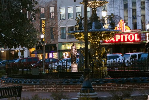 A decorative fountain in a park, with a theater marquee and buildings in the background, illuminated at dusk.