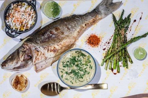 A whole grilled fish served with asparagus, creamy pasta, and garnishes on a branded tablecloth.