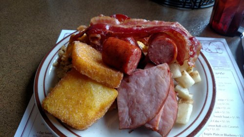 A hearty breakfast plate with bacon, sausage, ham, cornbread, and diced potatoes.