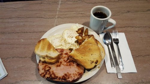 A plate with two fried eggs, toast, ham, and potatoes, accompanied by a cup of coffee and utensils on a table.