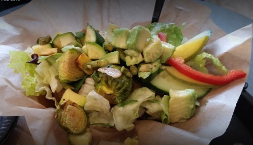 A fresh salad with Brussels sprouts, cucumbers, red pepper, and mixed greens in a paper bowl.