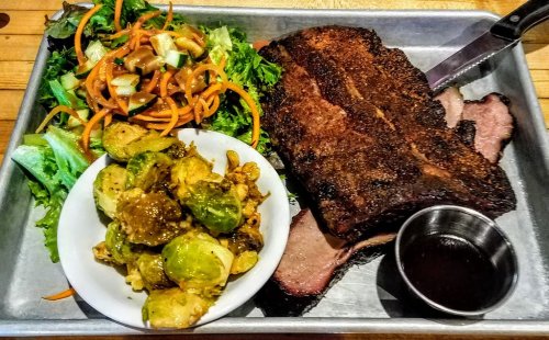 A metal tray with sliced smoked brisket, Brussels sprouts, and a fresh salad with carrots and cucumbers.