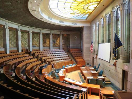 Interior of a legislative chamber with rows of seats, a large stained glass dome, and flags displayed.