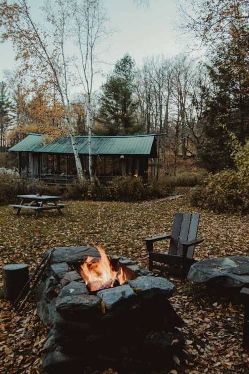 A cozy outdoor scene featuring a fire pit, wooden chair, and a rustic cabin surrounded by autumn foliage.
