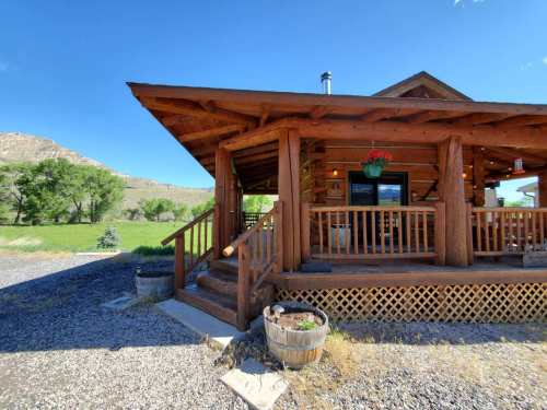 A rustic wooden cabin with a porch, surrounded by greenery and mountains under a clear blue sky.