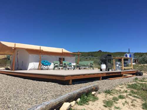 A glamping tent on a wooden deck, surrounded by gravel and greenery, with outdoor furniture and a grill under a clear blue sky.