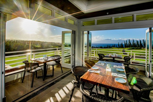 A bright restaurant with open doors, showcasing a scenic view of greenery and mountains under a sunny sky.