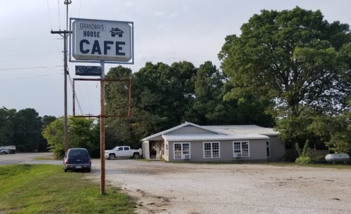 A roadside cafe named "Grandma's House" with a sign, surrounded by trees and a gravel parking area.