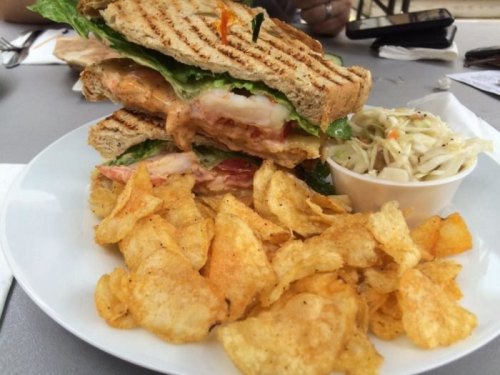 A stacked sandwich with shrimp, lettuce, and tomato, served with a side of potato chips and coleslaw.