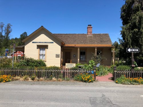 A charming yellow house with a porch, surrounded by flowers and greenery, on a sunny street with a one-way sign.