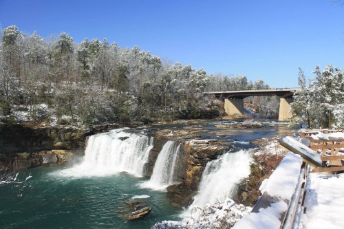 A snowy landscape featuring a waterfall and a bridge under a clear blue sky. Trees are dusted with snow.
