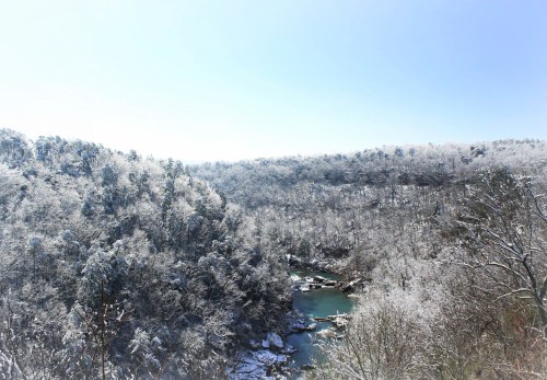 A snowy landscape with a river winding through a forested valley under a clear blue sky.