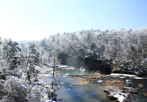 A serene winter landscape featuring a river surrounded by snow-covered trees and a clear blue sky.