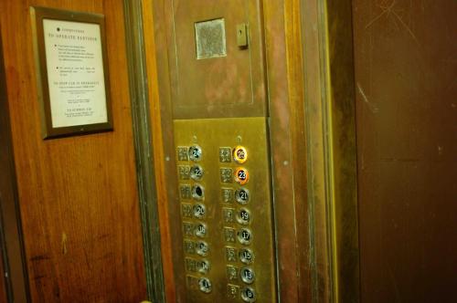 Vintage elevator panel with brass buttons, displaying floors 1 to 23, and a sign with instructions on the wall.