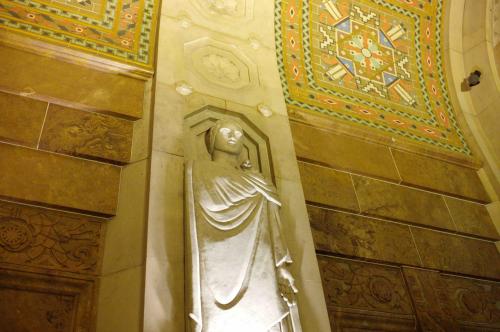 A stone statue of a figure draped in robes, set against a richly patterned wall with intricate designs.