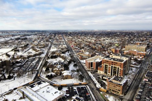 Aerial view of a snowy urban landscape with buildings, roads, and a distant waterfront under a cloudy sky.