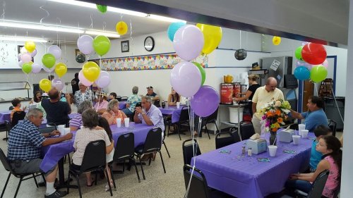 A festive gathering in a decorated hall with balloons, tables set for guests, and people socializing.