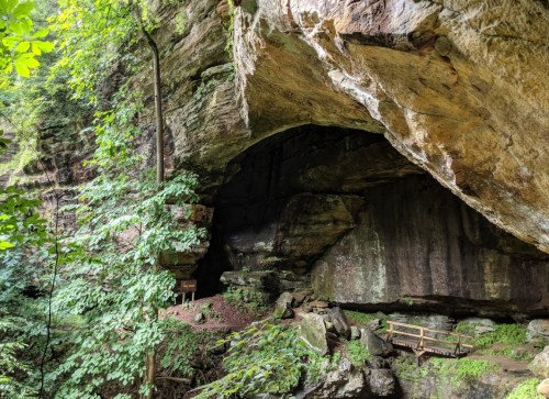 A rocky cave entrance surrounded by lush green trees and a wooden walkway in a natural setting.