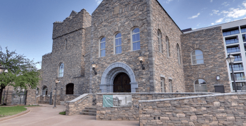 Stone building with arched entrance and large windows, surrounded by greenery and modern structures in the background.