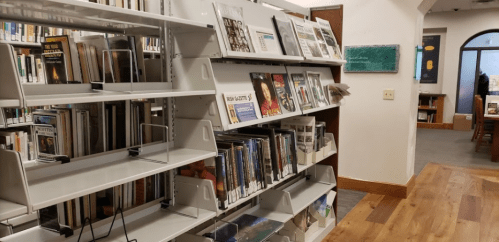 A library interior featuring bookshelves with books and magazines, wooden flooring, and a cozy reading area in the background.