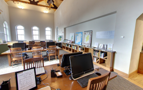 A spacious library with wooden desks, computers, and maps of Ireland on the walls, featuring large windows and bookshelves.