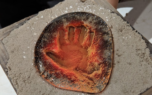 A handprint embedded in a translucent, fiery orange resin on a sandy surface.